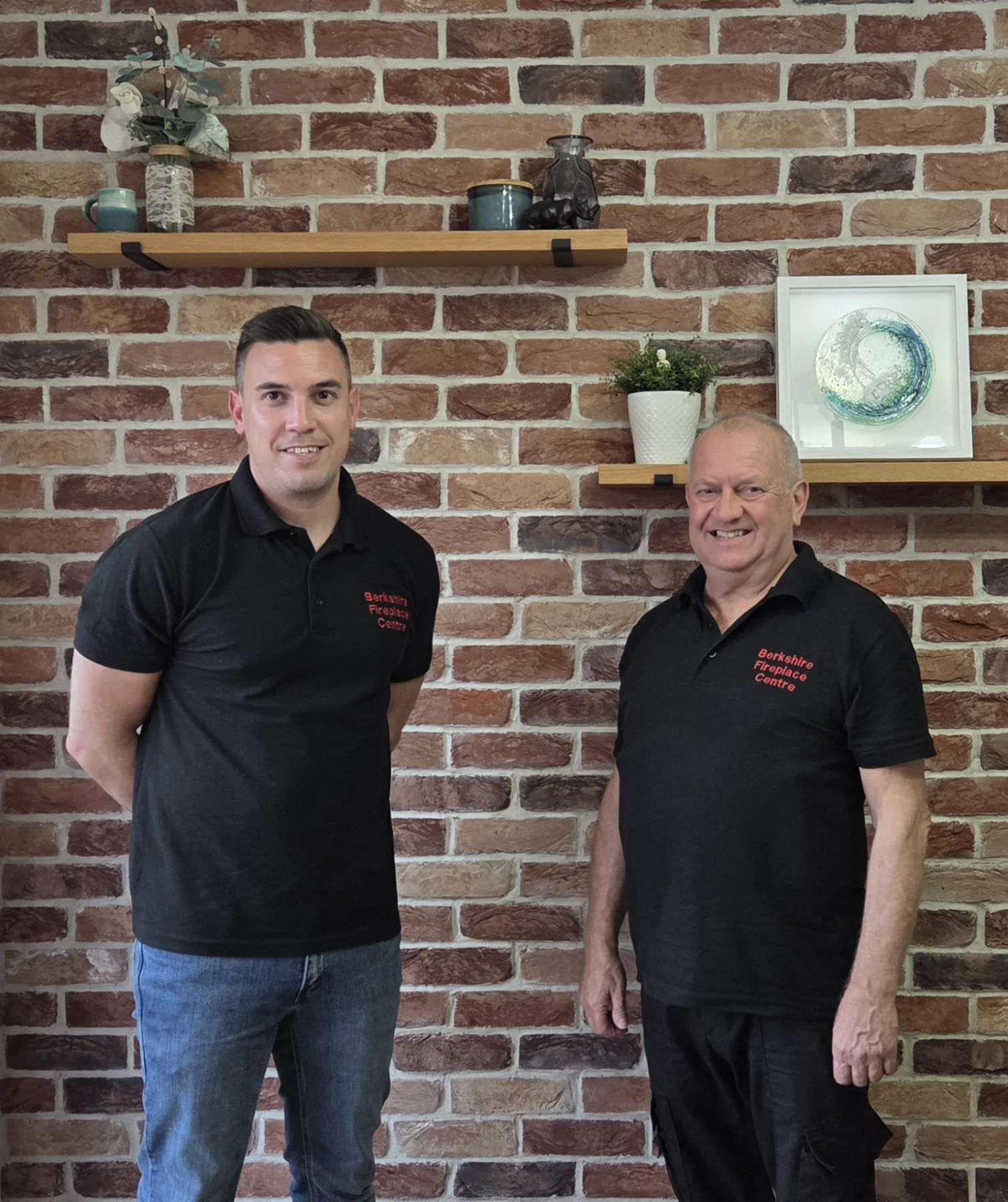 Graham and Matthew of Berkshire Fireplace Centre standing together in front of a brick wall, wearing their branded shirts.